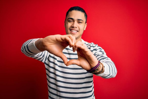 Young brazilian man wearing casual striped t-shirt standing over isolated red background smiling in love doing heart symbol shape with hands. Romantic concept.