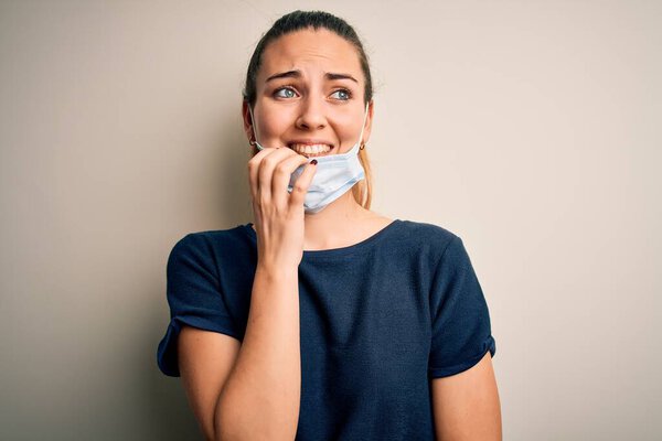 Beautiful blonde woman with blue eyes wearing medical mask over white background looking stressed and nervous with hands on mouth biting nails. Anxiety problem.