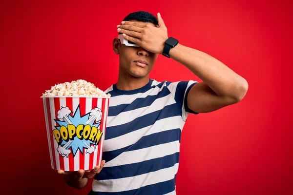 Young handsome african american man watching movie using 3d glasses eating popcorns covering eyes with hand, looking serious and sad. Sightless, hiding and rejection concept