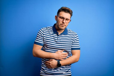 Mavi gözlü genç adam gözlük takıyor ve mavi arkaplanlı sıradan çizgili tişört giyiyor. Midesinde hazımsızlık, acı veren bir hastalık yüzünden. Ache kavramı.