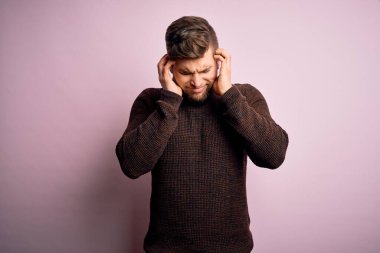 Sarışın, sakallı ve mavi gözlü genç bir adam pembe arka plan üzerine günlük süveter giyiyor ve baş ağrısı çekiyor çünkü stres yüzünden. Migren ağrısı..