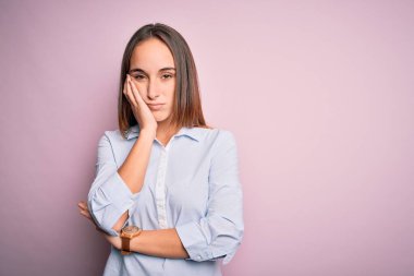 Zarif bir gömlek giyen genç ve güzel bir iş kadını pembe arka planda duruyor. Kollarını kavuşturup depresyondan sıkılmış ve yorgun görünüyor..
