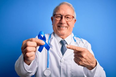 Kır saçlı kıdemli doktor elinde kolon kanseri farkındalığı mavi arka planda mavi kurdele çok mutlu bir el ve parmak işareti.