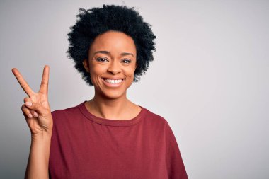 Kıvırcık saçlı, sıradan tişörtlü, genç ve güzel Afro-Amerikan kadın ayakta ve parmaklarıyla iki numarayı gösterirken kendinden emin ve mutlu bir şekilde gülümsüyor..