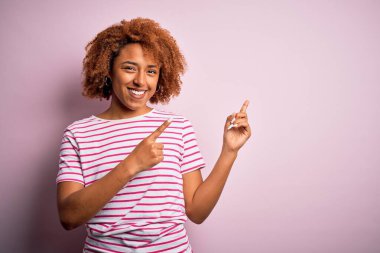 Kıvırcık saçlı, sıradan çizgili tişört giyen, gülümseyen ve kameraya bakan, iki eli ve parmağı yanda olan güzel Afro-Amerikan bir kadın..