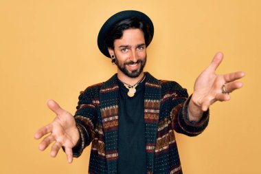 Genç, yakışıklı, bohem, hippi tarzı giyinmiş ve bohem şapkalı bir adam kameraya bakıp sarılmak için gülümsüyor. Mutluluğu kucaklayan neşeli bir ifade..