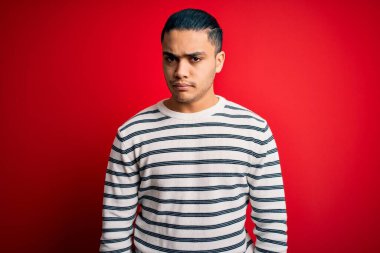 İzole edilmiş kırmızı arka plan üzerinde duran sıradan çizgili tişört giyen genç Brezilyalı bir adam şüpheci ve gergin, sorun yüzünden üzgün. Negatif kişi.