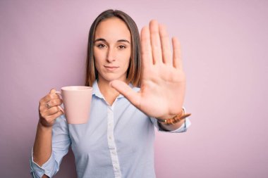 Genç, güzel bir kadın pembe arka planda durmuş kahve içiyor. Açık elle dur işareti yapıyor. Ciddi ve kendinden emin bir ifadeyle, savunma jesti yapıyor.