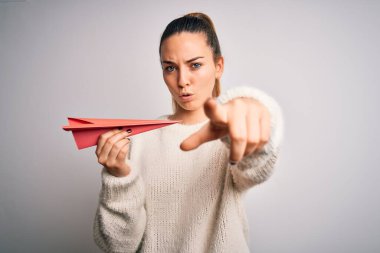 Mavi gözlü güzel sarışın kadın beyaz arka planda kırmızı kağıt uçak tutuyor. Kamerayı işaret ediyor. Ve sen, el işareti, ön taraftan pozitif ve kendinden emin bir hareket.