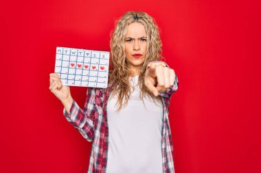 Genç, sarışın bir kadın adet takvimini tutuyor. Regl döngüsünü kontrol ediyor. Parmağıyla kamerayı işaret ediyor.