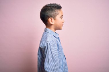 Zarif bir gömlek giyen küçük bir çocuk pembe izole edilmiş arka planda duruyor, yan yana bakıyor, rahat bir görünüm sergiliyor, kendine güvenen bir gülümsemeyle doğal bir yüz ifadesi sergiliyor..