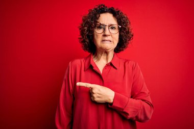 Orta yaşlı, kıvırcık saçlı, rahat gömlekli ve kırmızı arka planda gözlüklü güzel bir kadın. Endişeli ve gergin bir kenara işaret eden, endişeli ve şaşırmış bir ifade.