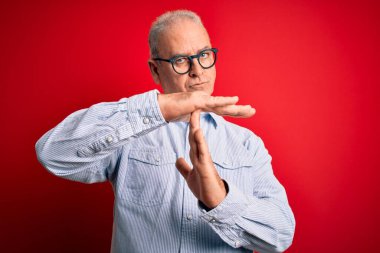 Orta yaşlı, yakışıklı, beyaz, sıradan çizgili gömlek ve kırmızı arka plan gözlüklü adam. Elleriyle jest yapıyor, hayal kırıklığına uğramış ve ciddi bir yüz ifadesi var.