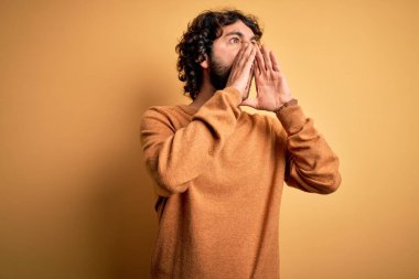 Sakallı, sıradan bir süveter giyen, sarı arka planda dikilen genç yakışıklı adam elleri ağzında sinirli bir şekilde bağırıyor.