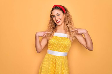 Sarı arka planda dikey duran mavi gözlü güzel sarışın kadın yüzünde bir gülümsemeyle gururlu ve mutlu birini işaret ediyor..