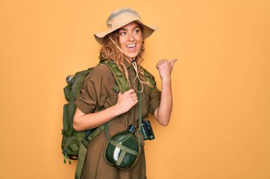 Mavi gözlü genç sarışın kaşif sırt çantası ve su matarası takmış gülümseyen ve başparmağıyla yan tarafı gösteren..