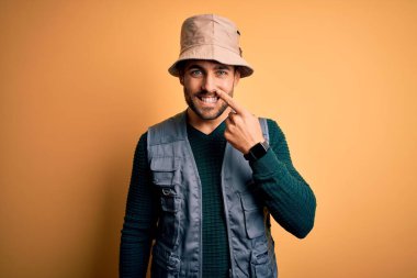 Tatilde sakallı, sarı arka planda kaşif şapkası takan, el ve burnu işaret eden, neşeli gülümseyen yakışıklı bir turist. Güzellik konsepti