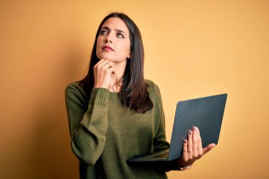 Mavi gözlü genç esmer kadın bilgisayarla çalışıyor sarı arka planda, elleri çenesinde soru sormayı, düşünceli ifadeyi düşünüyor. Düşünceli bir yüzle gülümsüyor. Şüphe kavramı.