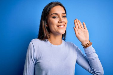 Genç, güzel, esmer bir kadın mavi arka planda dikilmiş, mutlu ve güler yüzlü, dostça bir hoş geldin jesti yapıyor.