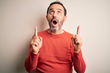 Orta yaşlı, turuncu süveter giyen, izole edilmiş beyaz arka planda dikilen beyaz adam şaşırmış ve şaşırmış, parmaklarıyla işaret ediyor ve kollarını kaldırıyordu..