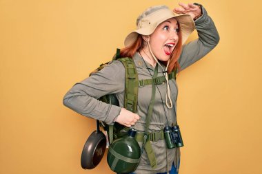 Sırt çantalı, sarı arka planda şapkalı, sırt çantalı, genç, kızıl saçlı kadın çok mutlu ve tebessüm ediyor. Konsept aranıyor.