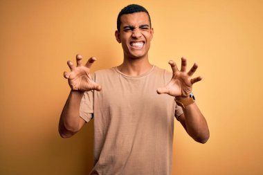 Genç, yakışıklı, Afro-Amerikalı bir adam sarı arka planda dikilen günlük tişört giyiyor komik bir şekilde kedi gibi pençe hareketi yapıyor, agresif ve seksi bir ifade.