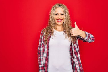 Genç, güzel, sarışın, günlük gömlekler giyen, izole edilmiş kırmızı arka planda duran, el hareketleriyle mutlu mesut hareket eden bir kadın. Kameraya bakıp başarıyı gösteren ifade onaylanıyor.