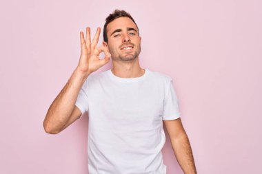 Mavi gözlü yakışıklı bir adam pembe arka planda duran günlük beyaz tişörtler giyiyor ve el ve parmaklarıyla 
