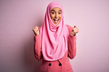 Pembe Müslüman başörtüsü ve iş ceketi giyen genç ve güzel esmer iş kadını kollarını kaldırarak ve açık gözlerle başarının şaşkınlığını ve şaşkınlığını kutluyor. Kazanan konsept.
