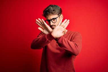 Sakallı, gözlüklü ve süveterli genç adam kırmızı arka planda duruyor.
