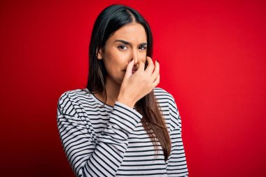 Genç, güzel esmer kadın kırmızı arka planda sıradan çizgili tişört giyiyor pis ve iğrenç bir koku alıyor, nefesini burnunda tutuyor. Kötü kokuyor.