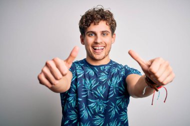 Sarışın, tatilde, kıvırcık saçlı, günlük yaz tişörtleri giyen, elle olumlu jest yapan, gülümseyen ve başarı için mutlu genç bir adam. Kazanan jest.