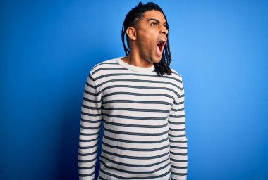 Young handsome african american afro man with dreadlocks wearing casual striped sweater angry and mad screaming frustrated and furious, shouting with anger. Rage and aggressive concept.