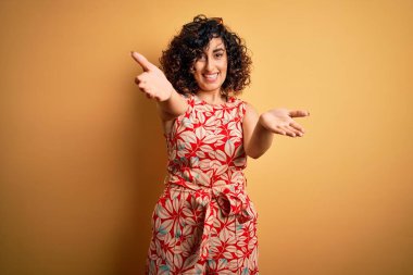 Genç, kıvırcık, güzel bir Arap kadın tatilde. Çiçekli bir elbise ve güneş gözlüğü takmış, kameraya bakıyor. Sarılmak için kollarını açmış gülümsüyor. Mutluluğu kucaklayan neşeli bir ifade..