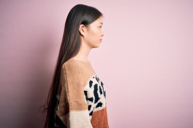 Hayvan desenli süveter giyen genç, güzel Asyalı bir kadın pembe izole edilmiş arka plana bakıyor, rahat profil pozu veriyor, kendine güvenen bir gülümsemesi var..