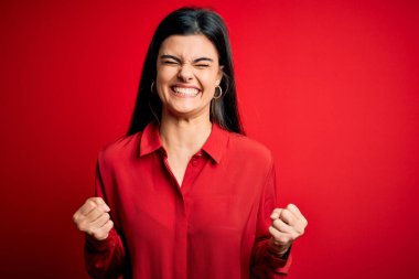Kırmızı arka planda dikilen rahat gömlekli genç, güzel esmer kadın. Başarı için heyecanlı kolları havada ve gözleri kapalı zaferi kutlarken gülümsüyor. Kazanan konsept.
