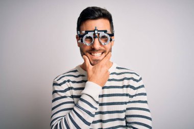 İzole edilmiş beyaz arka planda optometri gözlüğü takan sakallı yakışıklı genç adam çapraz kollar ve çeneyle gülümseyen kameraya bakıyor. Olumlu düşünüyorum..