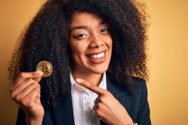 Afro saçlı, sarı arka planda bitcoin tutan Afro-Amerikalı genç bir iş kadını. Eli ve parmağı ile mutlu bir şekilde işaret ediyor.