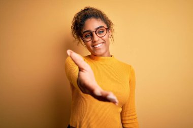 Sarı arka planda süveter ve gözlük takan güzel Afrikalı Amerikalı genç bir kız gülümseyen bir avuç içi yardım ve kabullenme eli uzatıyor..