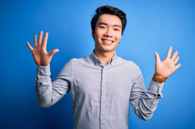 Genç, yakışıklı, sıradan bir gömlek giyen, mavi arka plan üzerinde duran ve parmaklarını on numara gösteren, kendinden emin ve mutlu gülümseyen Çinli bir adam..