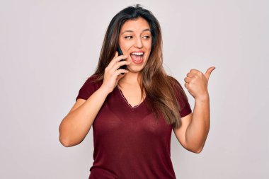 Genç İspanyol kadın akıllı telefondan konuşuyor izole bir arka plan üzerinden işaret ediyor ve gülümseyen bir yüzle baş parmağıyla gösteriyor.