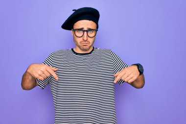 Mor arka plan üzerinde çizgili tişört ve Fransız beresi giyen yakışıklı adam üzgün ve üzgün görünüyor, parmaklarıyla yönü gösteriyor, mutsuz ve depresif..