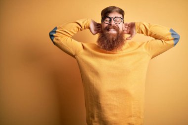 Sarıdan izole edilmiş arka planda gözlüklü yakışıklı kızıl saçlı, kolları ve elleri başının arkasında mutlu bir şekilde gülümseyen yakışıklı bir adam.