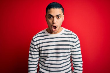 İzole edilmiş kırmızı arka planda dikilen sıradan çizgili tişört giyen genç Brezilyalı adam şaşkınlık ve şaşkınlık ifadesi, korku ve heyecan dolu yüz ifadesiyle dehşete düşmüş ve şok olmuştu..