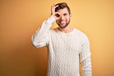 Sarışın, sakallı, mavi gözlü, sarı arka planda beyaz süveter giyen, el hareketleriyle gülümseyen, mutlu yüzüyle parmaklarının arasından bakan genç bir adam..