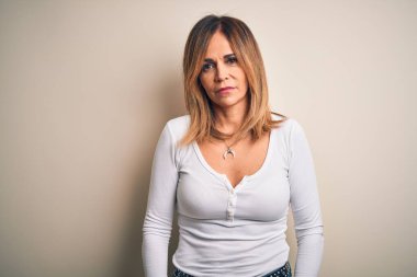 Yüzünde ciddi bir ifadeyle izole edilmiş beyaz arka planda duran günlük tişört giyen orta yaşlı güzel bir kadın. Basit ve doğal bir şekilde kameraya bakıyorsun..