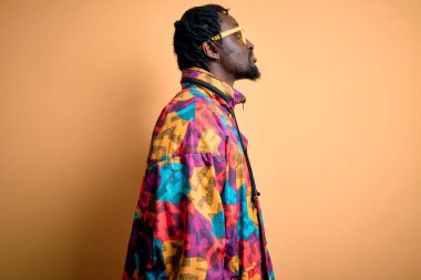 Sarı arka plan üzerinde gözlüklü, renkli ceketli, yakışıklı Afro-Amerikalı adam, rahat bir görünüm ve kendine güvenen bir gülümsemeyle poz veriyor..