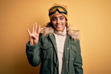 Genç Afrikalı Afro-Amerikan kayakçı kız kar spor giysisi ve kayak gözlüğü takıyor ve parmaklarıyla dört numarayı gösterirken kendinden emin ve mutlu bir şekilde gülümsüyor..