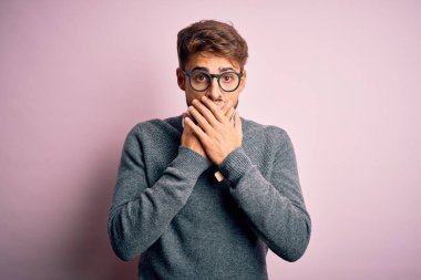 Gözlüklü, pembe arka planda duran sakallı ve kazaklı genç yakışıklı adam yanlışlıkla ağızlarını kapatarak şok geçirdi. Gizli konsept.