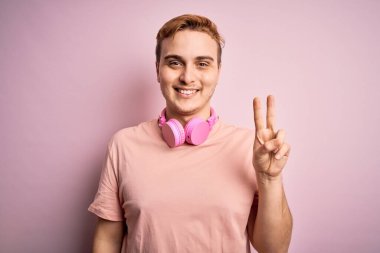 Genç, yakışıklı, kızıl saçlı, müzik dinleyen, pembe arka planda kulaklık takan, gülümseyen, kameraya göz kırpan, zafer işareti yapan bir adam. İki numara..